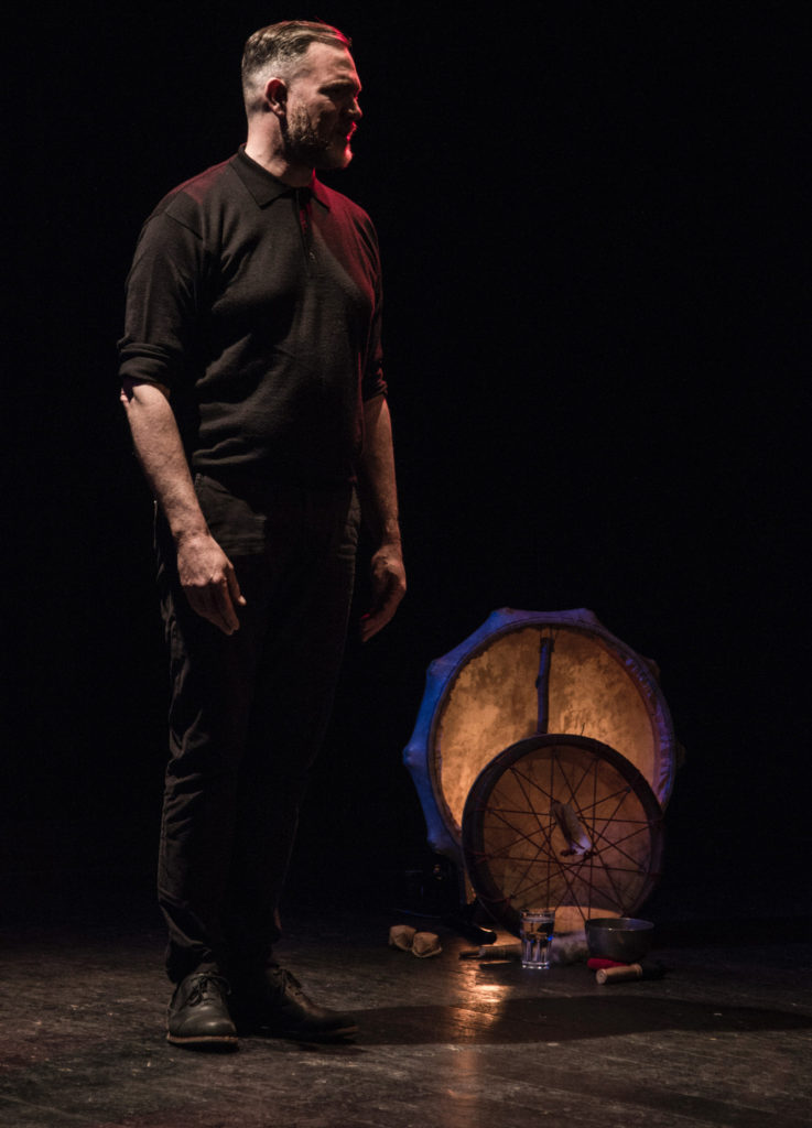 A male storyteller stands on stage during a live storytelling show at The Cube, Bristol. The man is dressed in black and the stage is dark.